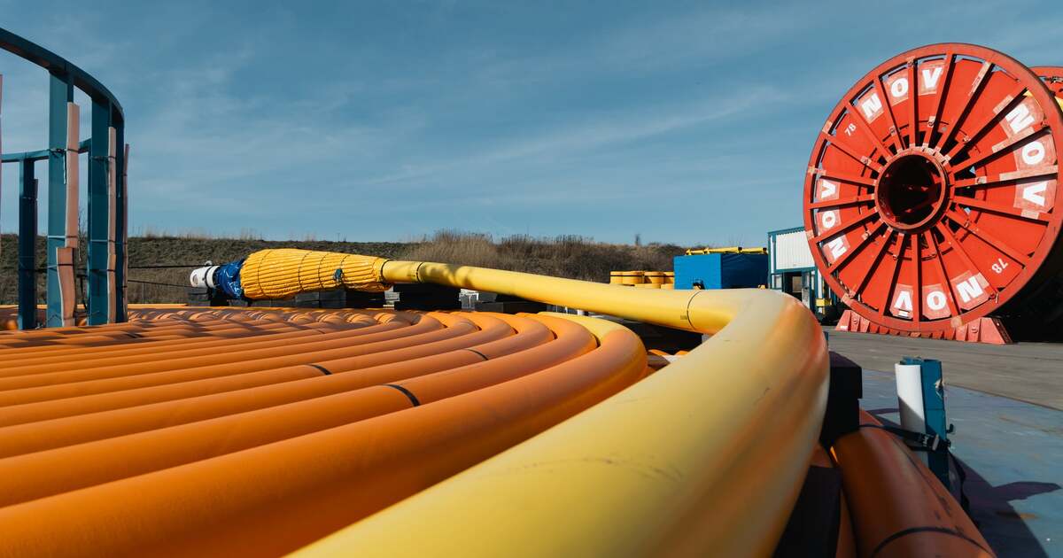 Large orange and yellow flexible offshore pipes and umbilicals are coiled in the foreground, leading toward a red industrial reel in an outdoor energy equipment storage yard under a clear blue sky.