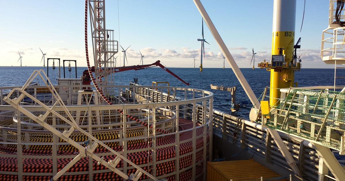 A large circular steel frame containing heavy duty undersea cabling alongside cable-laying equipment in the foreground and offshore wind turbines in the background